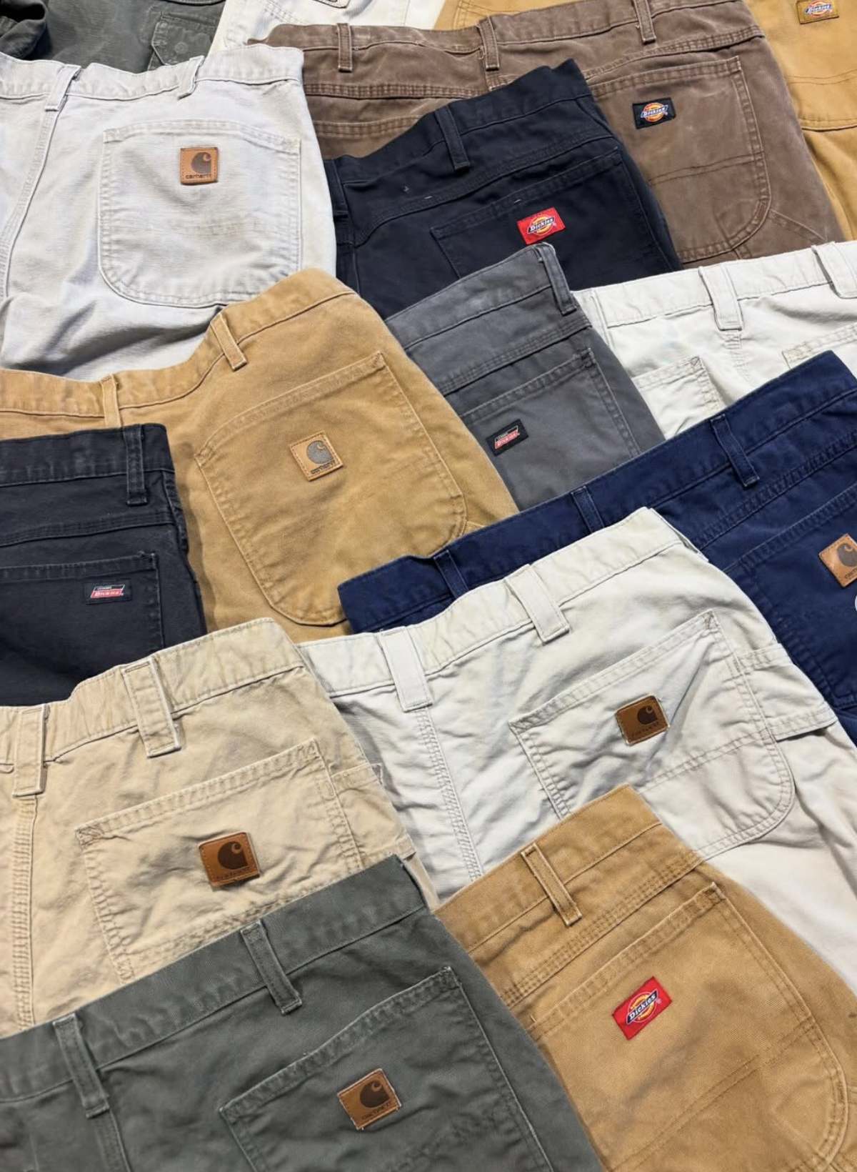Overhead view of a stack of folded Carhartt and Dickies carpenter shorts in tan, navy, gray, and cream.